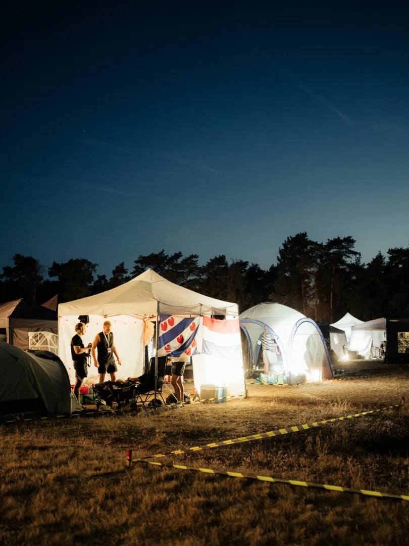 The base camp tents lit up at night in a forest clearing, a caution tape in front.
