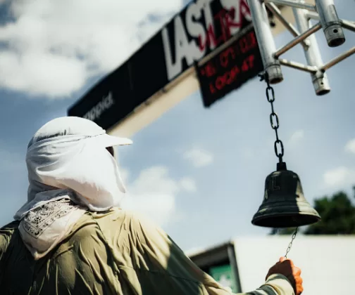 A runner in a white sun hood reaches for the start bell beneath the Last Soul race clock